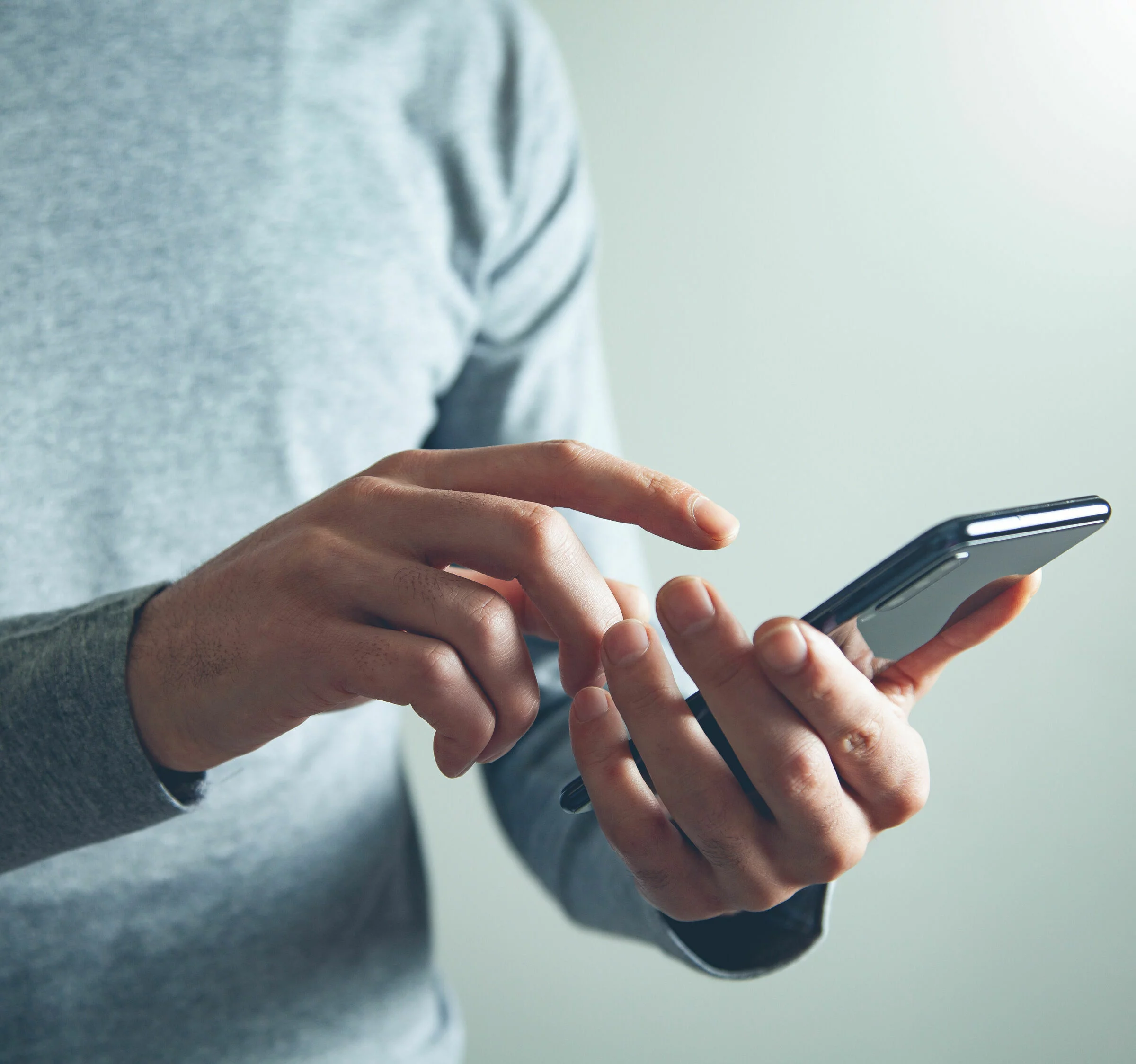 Person hält ein Smartphone in beiden Händen und tippt darauf. Die Person trägt ein graues Langarmshirt und befindet sich in einem hell erleuchteten Raum.